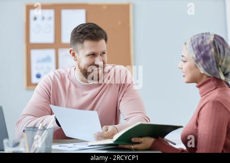 Happy young bearded businessman with paper looking at female colleague with smile while discussing points of financial document Stock Photo