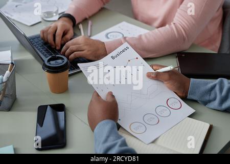 Financial document with graphic data held by young African American male broker sitting by workplace in front of colleague using laptop Stock Photo