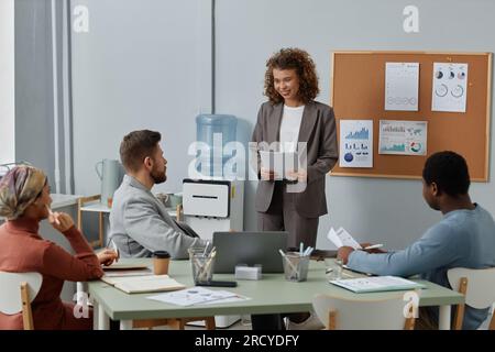 Young successful female broker with documents talking to one of colleagues sitting in front of her during discussion of working points Stock Photo