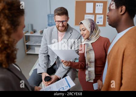 Two happy young male and female brokers looking at financial document at working meeting while sitting in front of intercultural colleagues Stock Photo