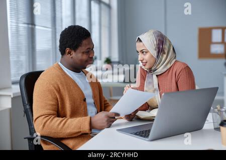 Young confident businesswoman in headscarf explaining financial data to African American male colleague at meeting while pointing at document Stock Photo