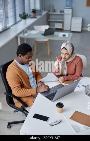 Above angle of two young intercultural colleagues in casualwear preparing presentation while sitting by workplace in front of laptop at meeting Stock Photo