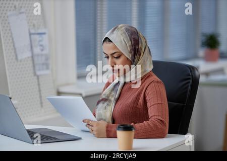 Young serious Muslim female entrepreneur or student in headscarf looking through document while sitting by workplace in front of laptop Stock Photo