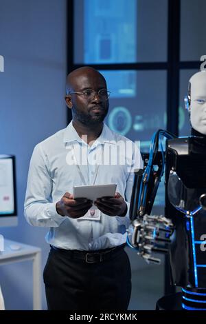 Vertical image of African American developer using tablet pc to control the robot in the lab Stock Photo