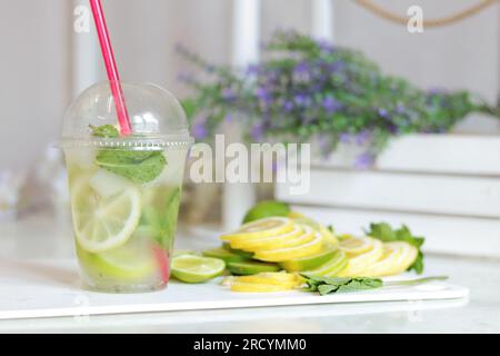 Making homemade mojitos. Close-up of a glass with ice, lemon, lime and mint. Lemonade preparation. A plastic cup with a mojito on a table in a light k Stock Photo