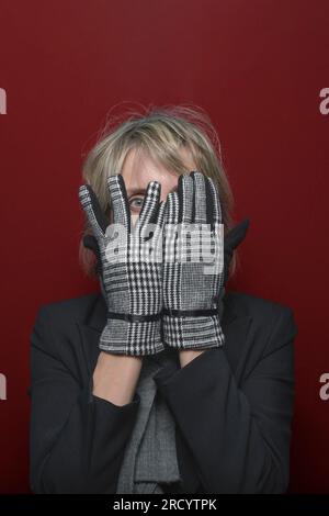 Turn a blind eye. A female model in elegant gloves looks through her fingers at the camera. portrait on a red background Stock Photo
