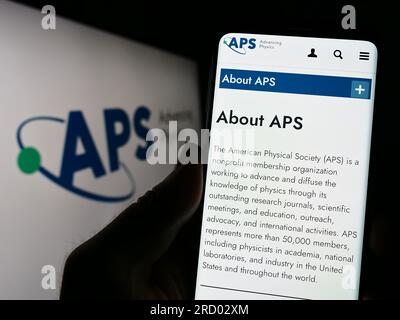 Person holding cellphone with webpage of US organization American Physical Society (APS) on screen with logo. Focus on center of phone display. Stock Photo