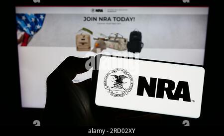 Person holding cellphone with logo of National Rifle Association of America (NRA) on screen in front of webpage. Focus on phone display. Stock Photo