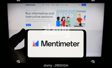 Person holding smartphone with logo of presentation software company Mentimeter on screen in front of website. Focus on phone display. Stock Photo