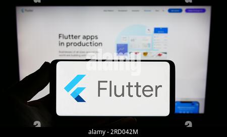Person holding smartphone with logo of UI software development kit Flutter (Google) on screen in front of website. Focus on phone display. Stock Photo