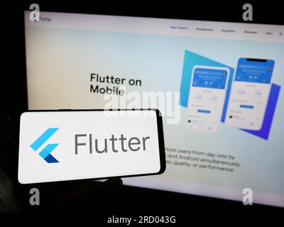 Person holding mobile phone with logo of UI software development kit Flutter (Google) on screen in front of web page. Focus on phone display. Stock Photo