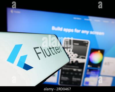 Mobile phone with logo of UI software development kit Flutter (Google) on screen in front of website. Focus on center-left of phone display. Stock Photo