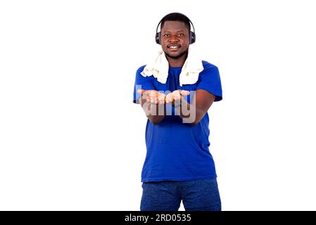 young man in sportswear standing in a gym with headphones and towel on his neck and showing his palms smiling. Stock Photo