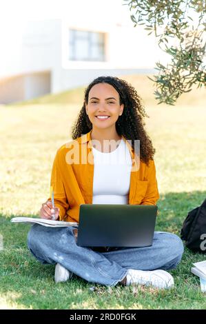 Learning concept. Vertical full-length photo of a stylish positive mixed race girl student, sitting on the grass near the tree, with a laptop and notepad, doing homework, taking notes, smile at camera Stock Photo