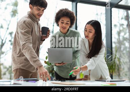 Three Young Multiracial Coworkers Pointing At Laptop Working Together Indoors Stock Photo