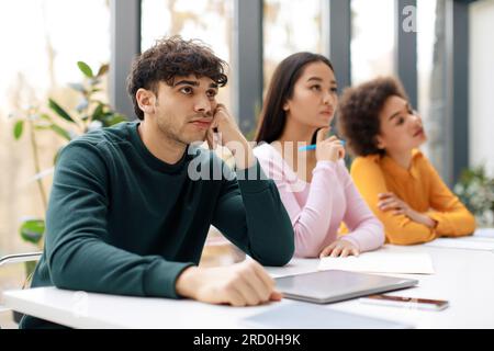 Bored european student guy leaning on hand and listening lecture, sitting with classmates at table in classroom Stock Photo