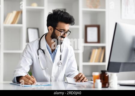 Handsome indian man doctor using computer and writing notes Stock Photo