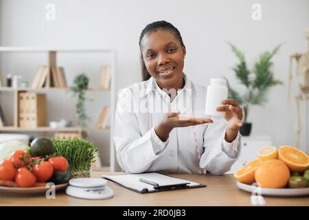 Cheerful multiracial woman offering pills container at camera during office hours in modern clinic. Smiling dietitian with measuring tape giving advice on supplements during weight loss program. Stock Photo