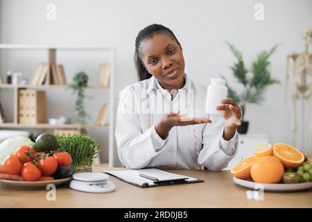 Cheerful multiracial woman offering pills container at camera during office hours in modern clinic. Smiling dietitian with measuring tape giving advice on supplements during weight loss program. Stock Photo