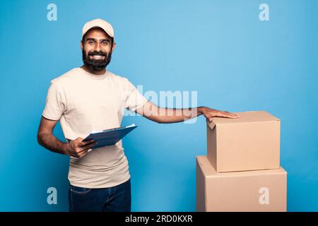 Delivery service concept. Happy indian courier man in uniform holding clipboard standing near cardboard boxes Stock Photo
