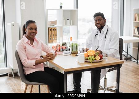 Full length portrait of multicultural people posing at office desk in doctor's workplace of medical center. Attractive woman in casual wear visiting professional dietitian on healthy eating issues. Stock Photo