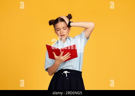 Tired unhappy teen european girl reads book, presses hand to head, isolated on yellow studio background Stock Photo