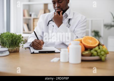 Expert in nutrition making special menu for patient and writing list of allowed products on clipboard paper during work time in hospital. African american man touching chin with hand and thinking. Stock Photo