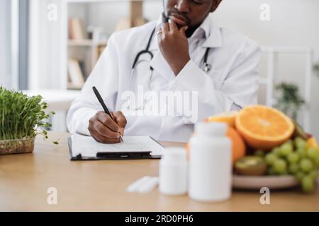 Expert in nutrition making special menu for patient and writing list of allowed products on clipboard paper during work time in hospital. African american man touching chin with hand and thinking. Stock Photo