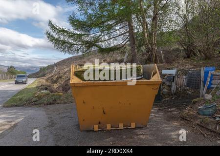 A bright yellow coloured skip with a mattresson the top which is covered in moss Stock Photo