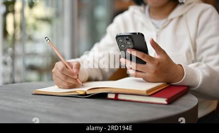 Cropped image of an Asian female college student using her smartphone while doing her homework at a cafe. Stock Photo