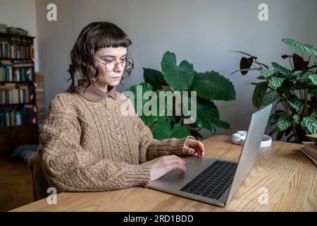 Focused teen student girl wearing glasses sitting at table typing on laptop Stock Photo