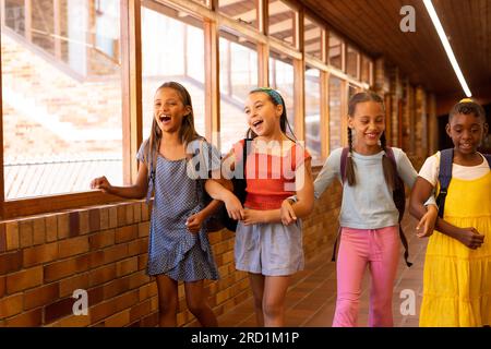 Diverse happy schoolgirls with bags walking and embracing in elementary school corridor Stock Photo
