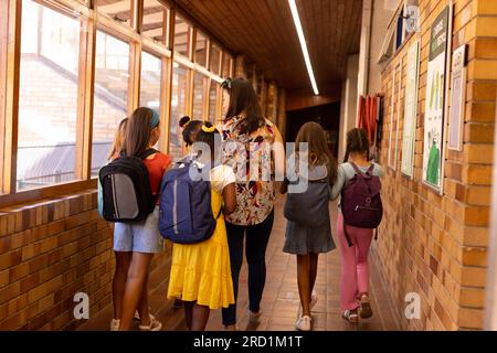 Diverse female teacher and schoolgirls with bags walking in elementary school corridor Stock Photo