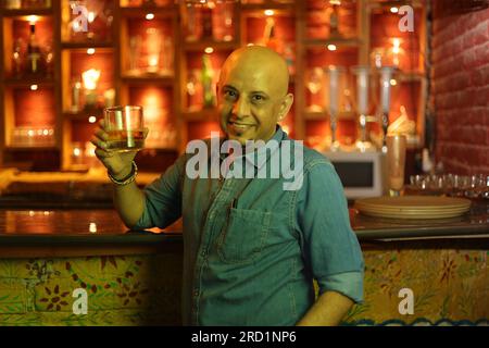 Young and flamboyant handsome bald guy standing on bar counter. Attractive man enjoying in city club interior at night club. Holding whiskey. Stock Photo