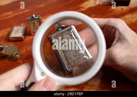 Using a magnifying glass to see the detail on an old Thai Buddha amulet Stock Photo