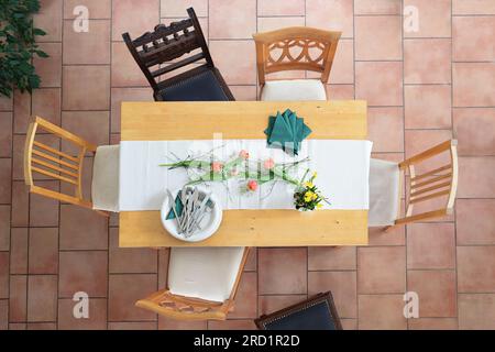 Preparation for a table setting with white plates and bowls, cutlery, green napkins and some tulip flowers, different wooden chairs around on a tiled Stock Photo