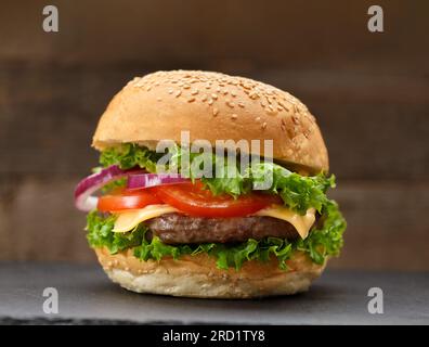 Appetizing burger with juicy beef cutlet, cheese, tomatoes, lettuce, onion on a black stone plate on a dark wooden background. Fast food Stock Photo