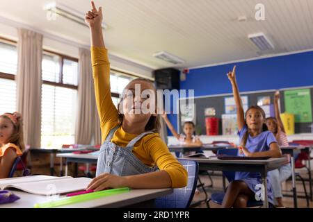 Happy diverse schoolgirls sitting at desks and raising hands in ...