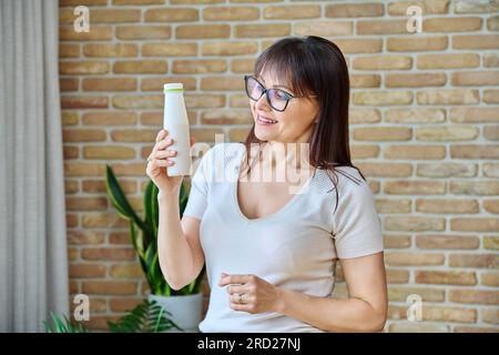 Middle aged woman drinking fermented milk product from bottle, near window Stock Photo