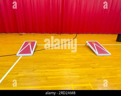 Front view of two homemade cornhole boards on a high school gym floor with a red curtain in the background. Stock Photo