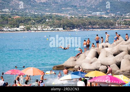 A youth dives into the sea from nearby rocks to cool down on a hot summer day. Others line up to follow him into the sea. Marbella, Spain Stock Photo
