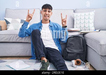 Young hispanic man sitting on the floor studying for university shouting with crazy expression doing rock symbol with hands up. music star. heavy musi Stock Photo