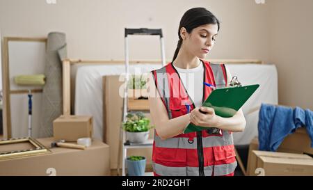 Young beautiful hispanic woman builder writing on clipboard at new home Stock Photo