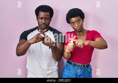 Young african american couple standing over pink background rejection expression crossing fingers doing negative sign Stock Photo