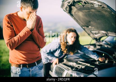 A young couple had a car breakdown on the road. They stand very upset, open the hood and try to fix the broken engine Stock Photo