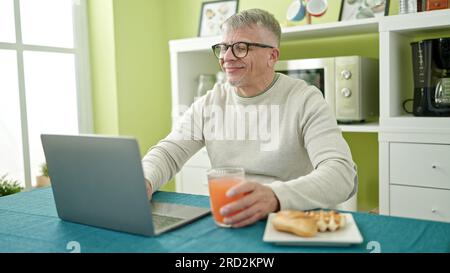 Middle age grey-haired man having breakfast using laptop at home Stock Photo