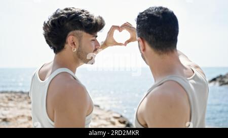 Two men couple hugging each other doing heart gesture at seaside Stock Photo