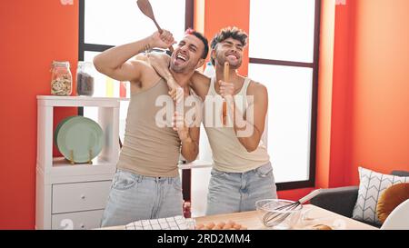 Two men couple singing song using kitchen utensil as a microphone at dinning room Stock Photo