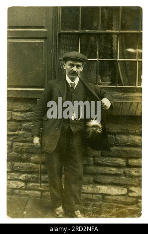 Original, charming early 920's postcard of working class man with cute toy or small Yorkshire terrier dog or puppy inside coat pocket outside a building possibly a pub. He wears a  Maltese Cross on a watch fob chain, possibly a WW1 veteran or fireman. Possibly Yorkshire, England, U.K. Stock Photo