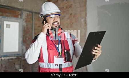Young latin man builder talking on smartphone reading document at construction site Stock Photo